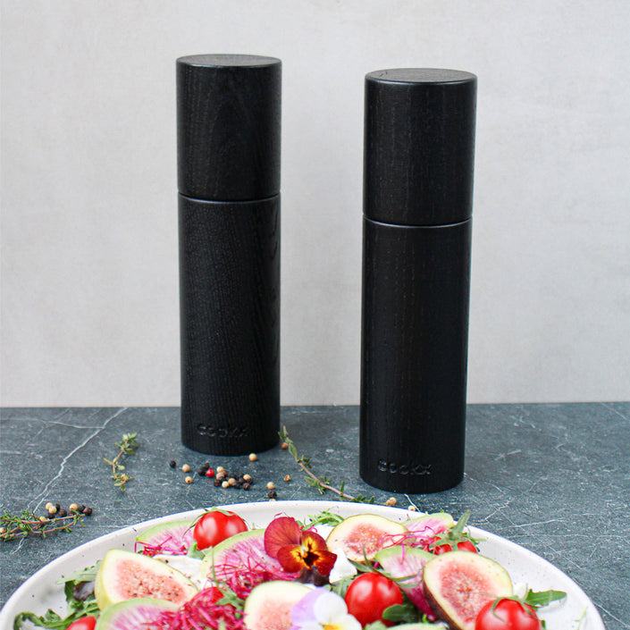 Salz-/Pfeffermühle Set aus Eichenholz schwarz, 23cm hoch, Milieubild in Küche, Set steht hinter Teller mit Salat. Frontalansicht