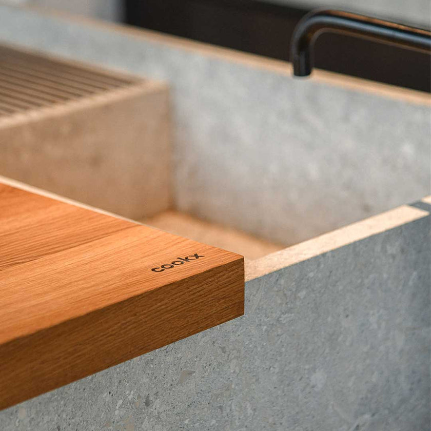 Hackbrett aus Eichenholz, Antirutschkante, hochkant 34 x 50 x 3,5 cm, rechts unten Logo von COOKX eingraviert, Milieubild in Küche, Brett liegt neben Spülbecken, Ansicht auf eingraviertes Logo