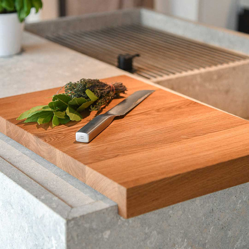 Hackbrett aus Eichenholz, Antirutschkante, hochkant 34 x 50 x 3,5 cm, rechts unten Logo von COOKX eingraviert, Milieubild in Küche, Brett liegt auf Kücheninsel, Kräuter und silbernes Messer auf Brett