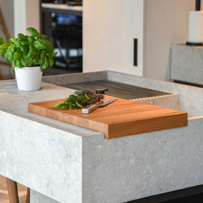 Hackbrett aus Eichenholz, Antirutschkante, hochkant 34 x 50 x 3,5 cm, rechts unten Logo von COOKX eingraviert, Milieubild in Küche, Brett liegt auf Kücheninsel, Kräuter und silbernes Messer auf Brett