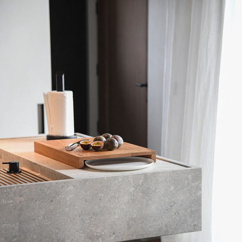 Hackbrett aus Holz Natur, Auffangöffnung für Teller, Milieubild in Küche, weißer Teller in Auffangöffnung, Messer und Früchte liegen auf Brett