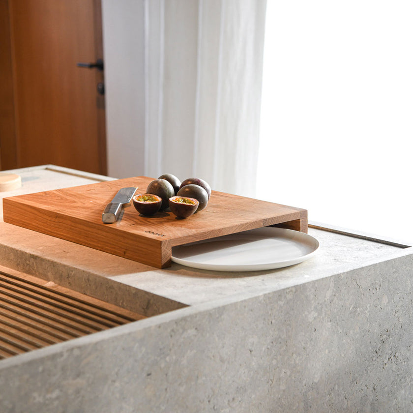 Hackbrett aus Holz Natur, Auffangöffnung für Teller, Milieubild in Küche, weißer Teller in Auffangöffnung, Messer und Früchte liegen auf Brett, Seitenansicht schräg 