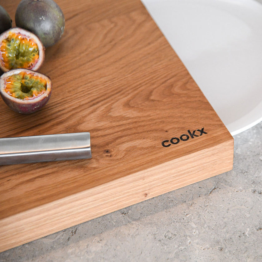 Hackbrett aus Holz Natur, Auffangöffnung für Teller, Milieubild in Küche, weißer Teller in Auffangöffnung, Messer und Früchte liegen auf Brett, Ecke mit Cookx Aufschrift 