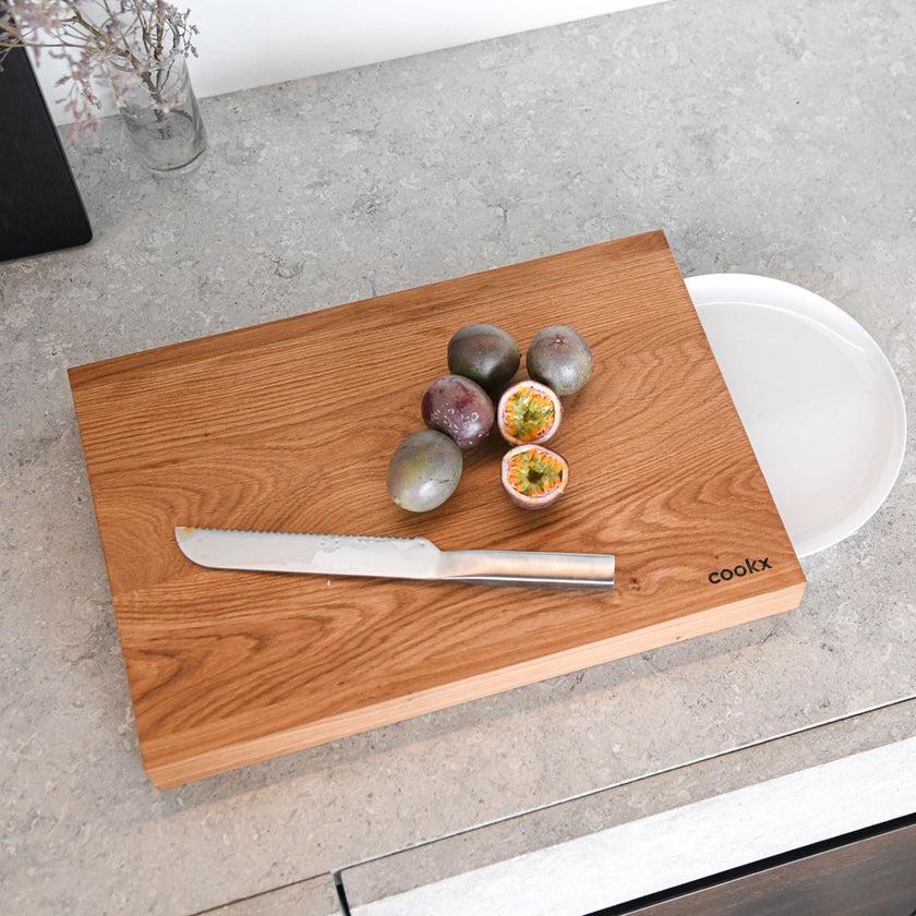 Hackbrett aus Holz Natur, Auffangöffnung für Teller, Milieubild in Küche, weißer Teller in Auffangöffnung, Messer und Früchte liegen auf Brett, Ansicht von oben