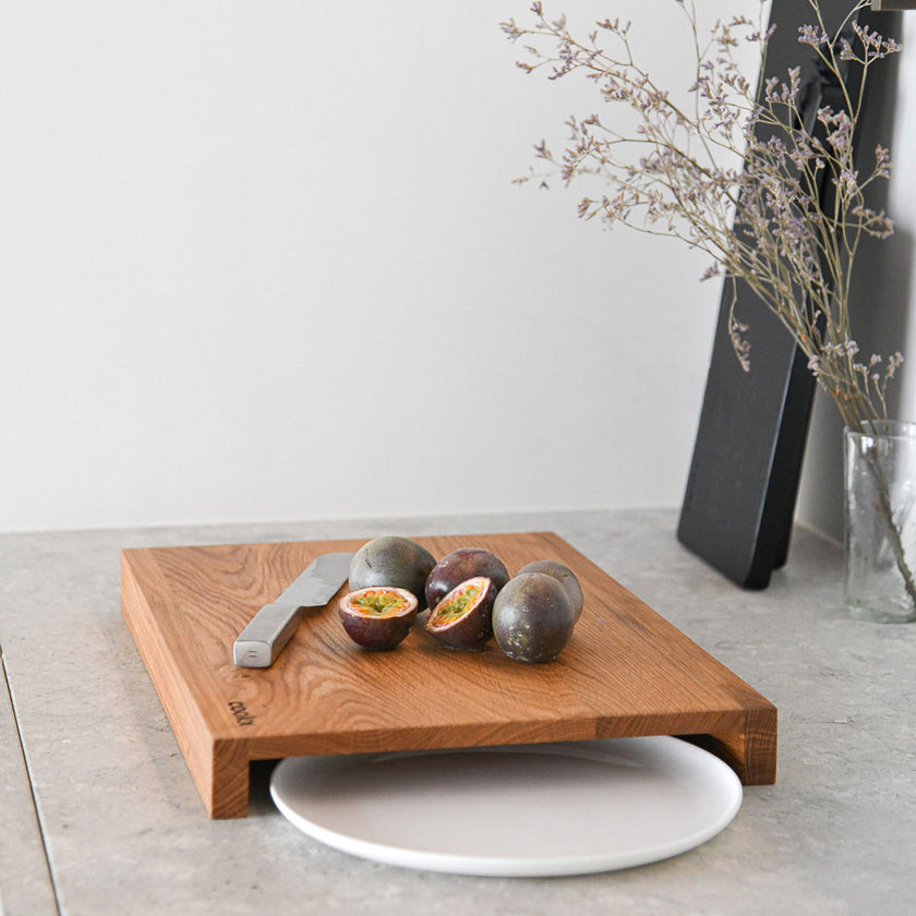 Hackbrett aus Holz Natur, Auffangöffnung für Teller, Milieubild in Küche, weißer Teller in Auffangöffnung, Messer und Früchte liegen auf Brett, Seitenansicht