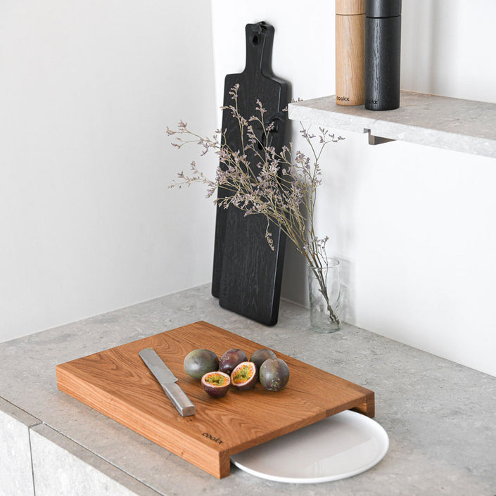 Hackbrett aus Holz Natur, Auffangöffnung für Teller, Milieubild in Küche, weißer Teller in Auffangöffnung, Messer und Früchte liegen auf Brett