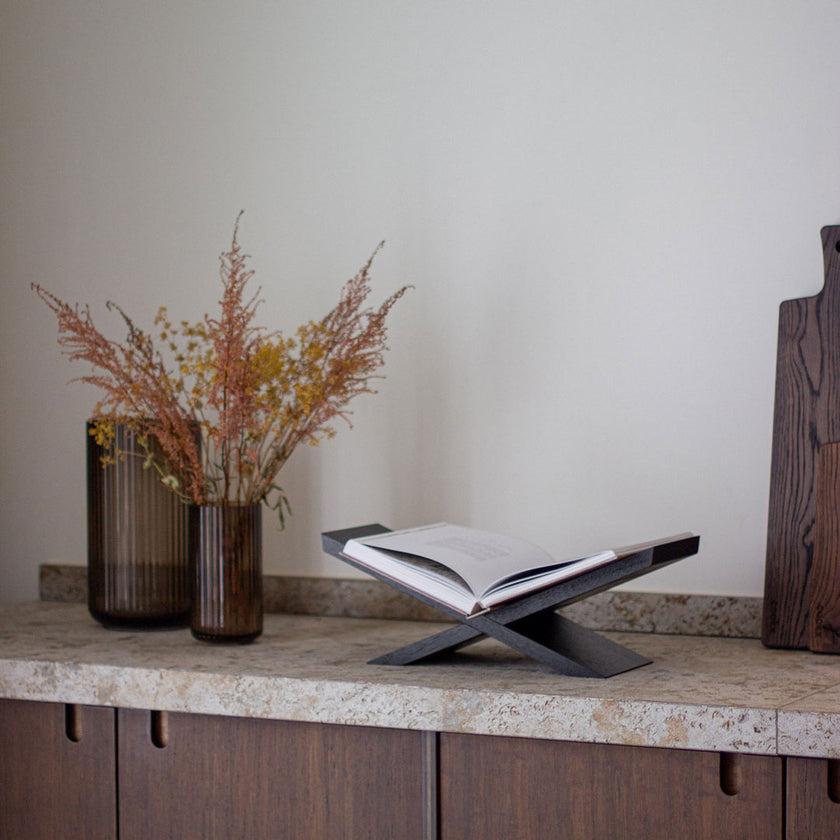 Buchständer aus Eichenholz schwarz, x-From, aus zwei gekreuzten Holzelementen, Milieubild frontal auf Arbeitsfläche in der Küche mit Buch