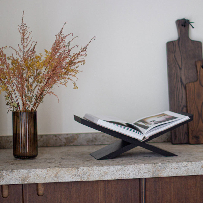 Buchständer aus Eichenholz schwarz, x-From, aus zwei gekreuzten Holzelementen, Milieubild frontal auf Arbeitsfläche in der Küche mit Buch