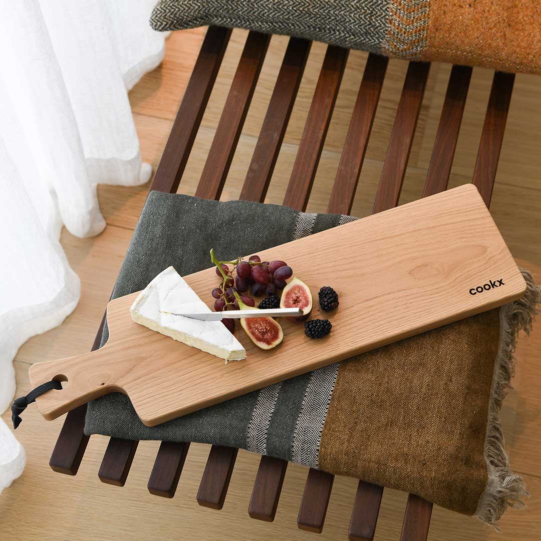 Servierbrett oder Schneidebrett aus Eichenholz Natur mit Griff und schwarzem Lederband zum aufhängen, 800mm x 18mm x 25mm, Milieubild im Wohnzimmer auf Bank, Brett mit Käse und Früchten liegt auf Decke 
