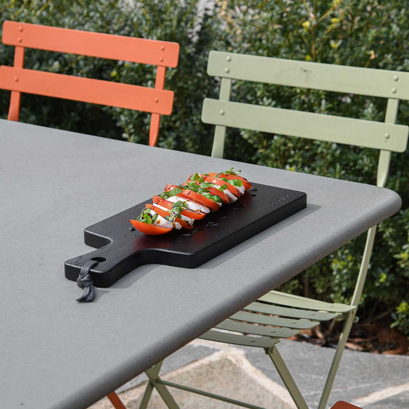 Servierbrett oder Schneidebrett aus Eichenholz schwarz mit Griff und schwarzem Lederband zum aufhängen, 400mmx180mmx25mm, Milieubild im Garten auf Gartentisch, Brett mit Tomate Mozzarella drauf