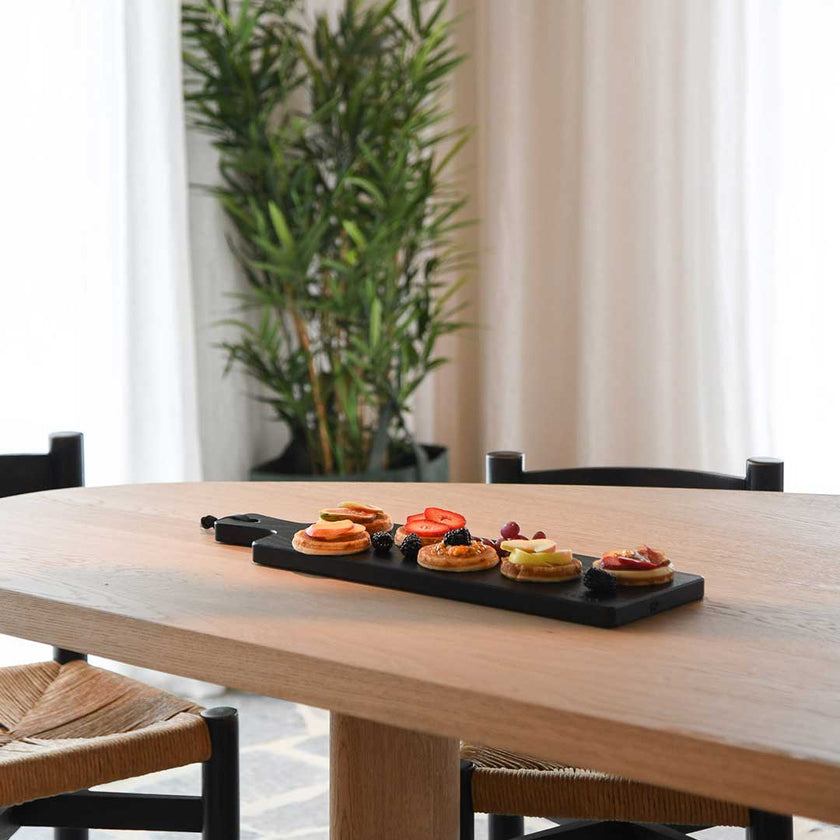 Servierbrett oder Schneidebrett aus Eichenholz schwarz mit Griff und schwarzem Lederband zum aufhängen, 600mmx180mmx25mm, Milieubild im Esszimmer auf Tisch, Brett mit Essen drauf