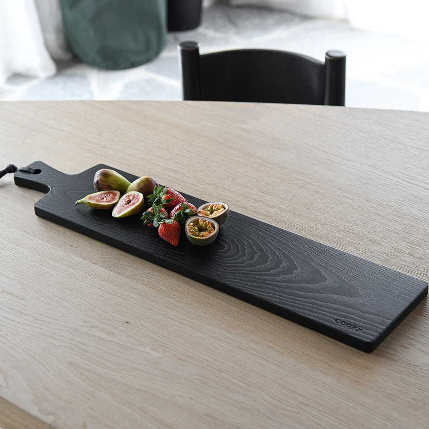 Servierbrett oder Schneidebrett aus Eichenholz schwarz mit Griff und schwarzem Lederband zum aufhängen, 800mmx180mmx25mm, Milieubild im Esszimmer auf Tisch, Brett mit Essen drauf