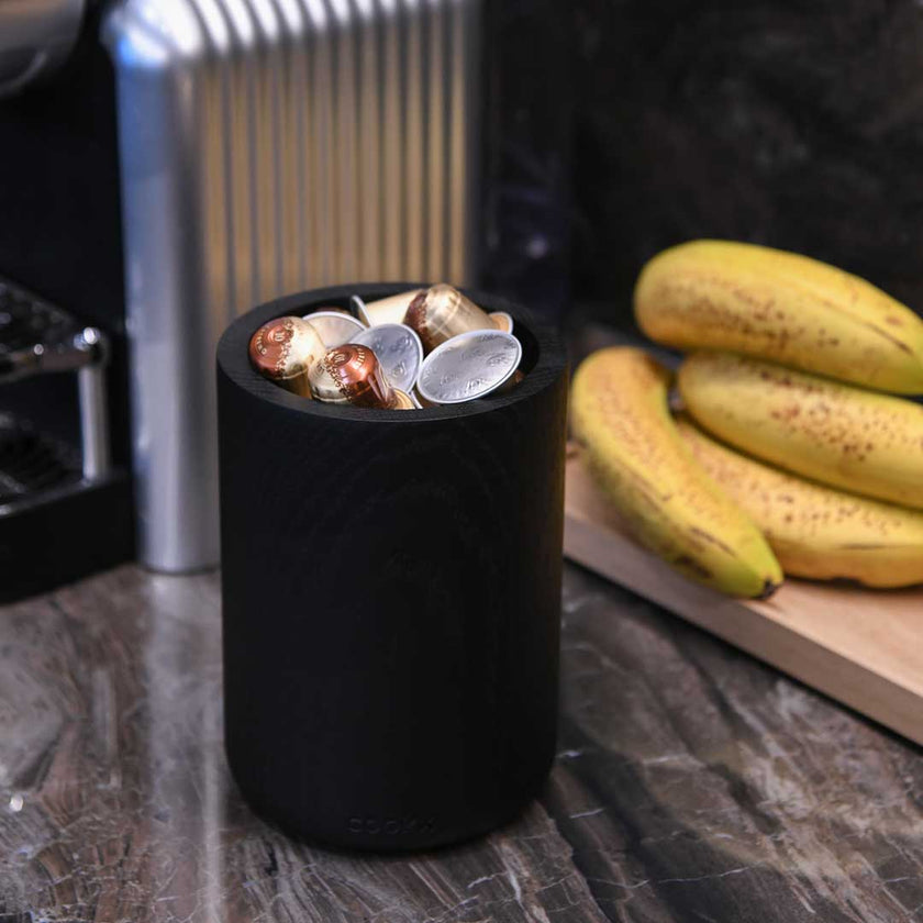 Küchenutensilienhalter aus Eichenholz schwarz, Milieubild in Küche, im Halter sind Kaffeekapseln und im Hintergrund liegt ein Bund Bananen 