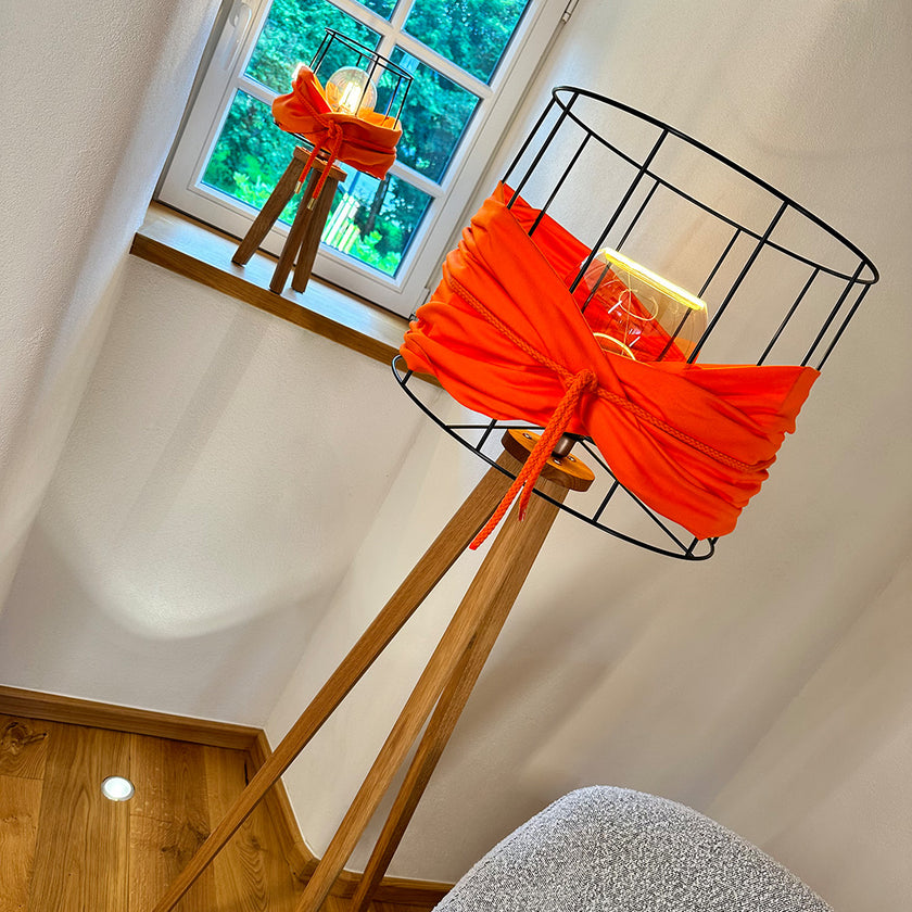 Stehleuchte Asteiche geölt, Modell Lore, Lampengestell offener schwarzer Metallrahmen, orangener Stoff um Metallrahmen, orangene Kordel um Stoff, Stromkabel verläuft durch ein Bein, Milieubild im Showroom, gleiche Leuchte als Tischleuchte auf Fensterbank