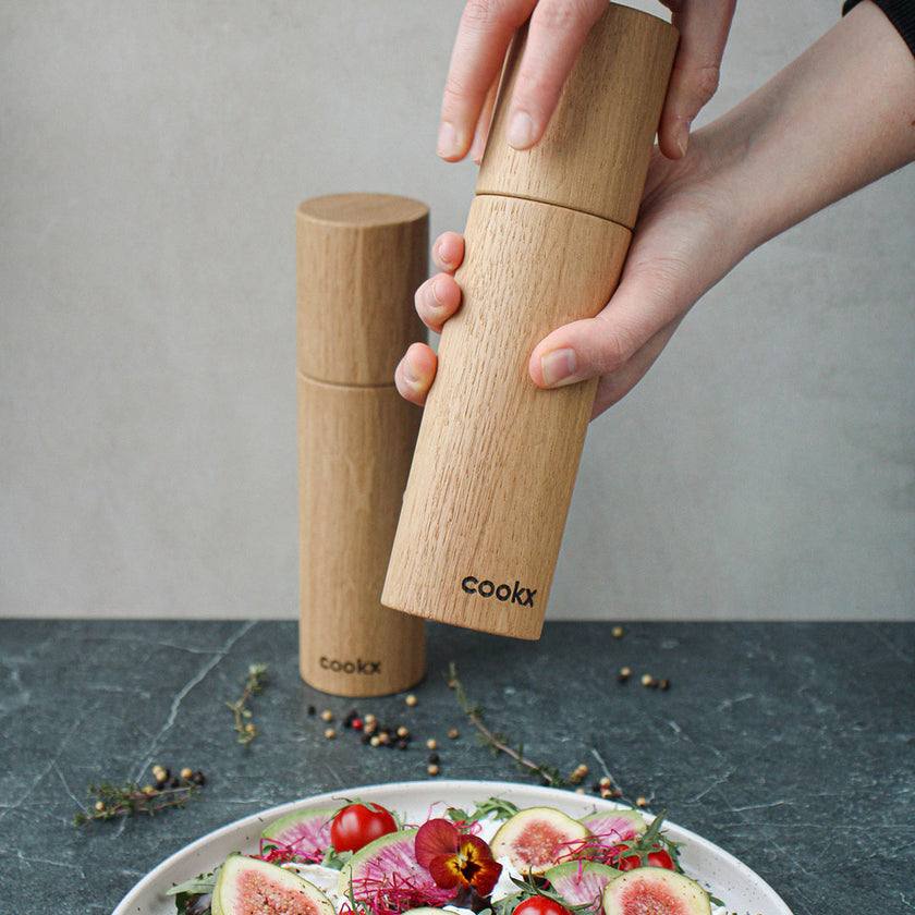 Salz-/Pfeffermühle Set aus Eichenholz Natur, 23cm hoch, Milieubild in Küche, Set steht hinter Teller mit Salat, eine Mühle wird benutzt, Frontalansicht