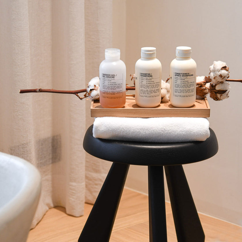 Kerzentablett aus Eichenholz Natur,  28 x 14 x 2,3 cm, Milieubild in Badezimmer, Tablett mit 3 Flaschen Shampoo steht auf schwarzem Hocker mit weißem Handtuch